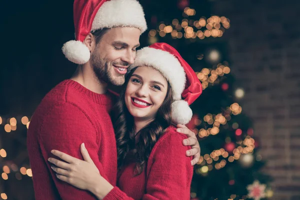 Foto de close-up do casal feliz passar véspera de Natal tempo na sala de luzes decoradas guirlanda perto de árvore de x-mas dentro de casa vestindo pulôveres vermelhos e chapéus de Papai Noel — Fotografia de Stock