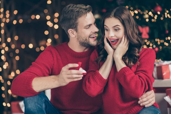 Foto de pareja en adornado guirnalda luces habitación chico dando señora inesperada participar anillo caja espera respuesta sentado acogedor cerca de árbol de x-mas en el interior usar suéteres rojos —  Fotos de Stock