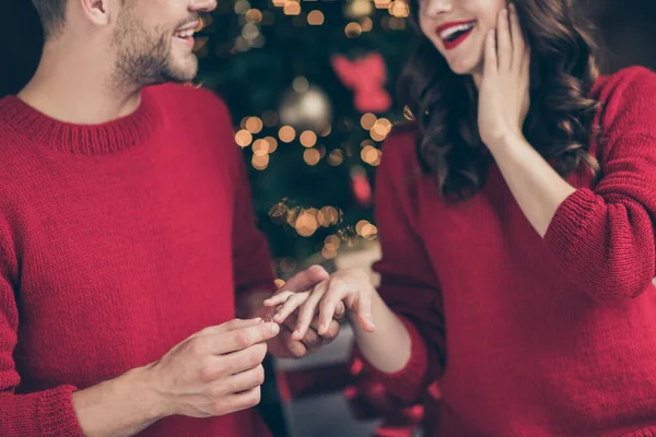 Will you marry me. Cropped photo of couple in love guy making proposal in decorated garland room near x-mas tree putting golden ring on lady finger indoors wear red sweaters