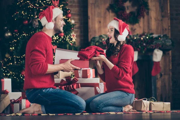 Foto van dromerig paar in versierde slinger lichtkamer uitwisselen vakantie geschenken zitten gezellig op de vloer in de buurt van x-mas boom binnen dragen rode truien en santa hoeden — Stockfoto