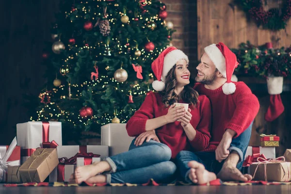 Foto de casal sonhador noite de ano novo em luzes decoradas guirlanda quarto sentado aconchegante no chão perto de árvore de x-mas beber bebida quente dentro de casa vestindo camisolas vermelhas e chapéus de santa — Fotografia de Stock