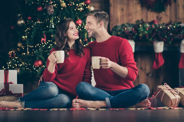 Foto von niedlichen Paar verbringen Neujahr Nacht in dekorierten Girlanden Lichter Zimmer sitzt gemütlich auf dem Boden in der Nähe immergrünen Baum trinken Heißgetränk drinnen tragen rote Pullover — Stockfoto