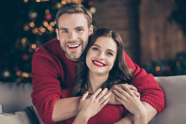 Closeup foto de casal bonito se preparando para a véspera de Natal na sala de luzes decoradas guirlanda perto de árvore do ano novo piggyback sentado sofá acolhedor dentro de casa vestindo camisolas vermelhas — Fotografia de Stock