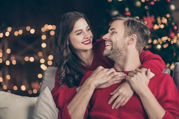 Foto de pareja encantadora pasando la víspera de Navidad en la sala de luces de guirnalda decorada abrazando a cuestas cerca de un árbol de año nuevo sentado acogedor sofá interior usar pullovers rojos —  Fotos de Stock