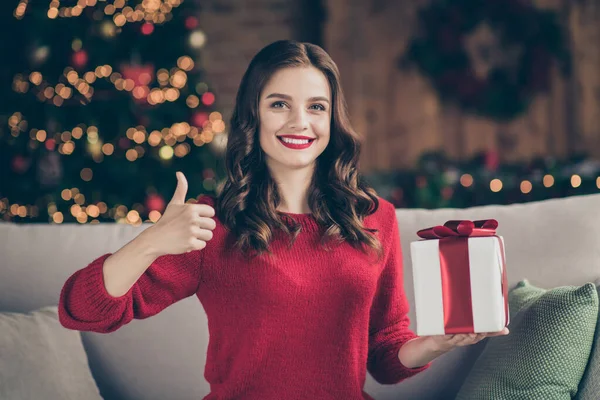 Foto de la señora divertida cogida de la mano caja de regalo asesorar a los compradores las ventas de año nuevo levantando el pulgar hacia arriba sentado sofá en la sala de luces de guirnalda decorada en interiores con pullovers rojos —  Fotos de Stock