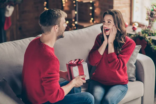 Foto de una linda pareja emocionada compartiendo regalos en víspera de x-mas pasando tiempo en la sala de luces de guirnalda decorada sentada acogedora en el sofá interior con pulóveres rojos —  Fotos de Stock