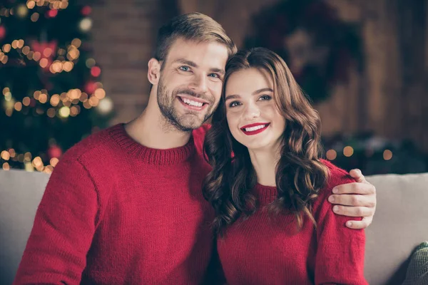 Foto de primer plano de pareja muy alegre pasar la víspera de Navidad de acebo en la sala de luces de guirnalda decorada sentado cerca de interiores con pullovers rojos —  Fotos de Stock
