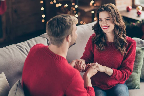 Za widokiem zdjęcie emocjonalnej pary trzymającej ramiona spędzające Boże Narodzenie razem w pokoju z lampkami do pończoch siedząc wygodnie na kanapie wewnątrz w czerwonych swetrach — Zdjęcie stockowe