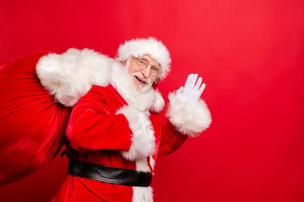 Portret van hem hij mooi vrolijk vrolijk baard Kerstman dragen bont jas dragen leveren zak hulst vrolijk ho-ho-ho zwaaien hi goedenacht geïsoleerd over helder levendig glans levendig rood achtergrond — Stockfoto