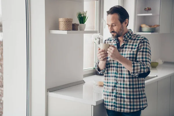 Foto de alegre optimista descansando guapo agradable contento en buen humor chico sosteniendo taza de delicioso café en las manos de pie cerca de la ventana grande — Foto de Stock