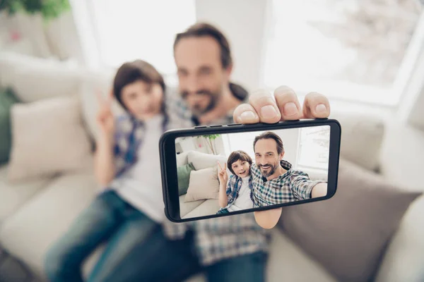 Phto retrato de guapo padre alegre positivo abrazando abrazando a su hijo en el sofá tomando selfie en el teléfono mostrando v-signo de tener mejor fin de semana sentado en la sala de estar en el interior —  Fotos de Stock