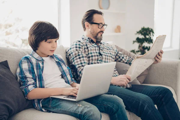 在明亮的白色现代室内客厅里，两个漂亮而又集中注意力的男人，爸爸和十多岁的儿子坐在沙发上，在笔记本电脑上打字，阅读新闻 — 图库照片