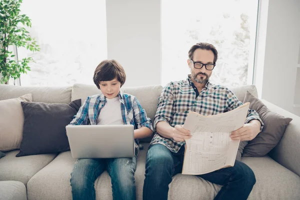 Retrato de dos simpáticos y atractivos chicos concentrados lindos y lindos papá e hijo pre-adolescente sentados en el sofá usando el ordenador portátil noticias de lectura en blanco claro estilo moderno casa interior sala de estar —  Fotos de Stock