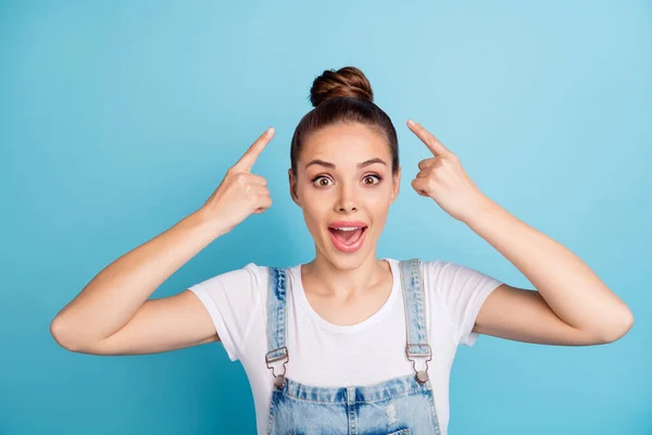 Porträt eines beeindruckten Mädchens, das mit erhobenem Zeigefinger auf ihren Haarschnitt zeigt und weißes T-Shirt Jeans Overalls trägt, die isoliert vor blauem Hintergrund stehen — Stockfoto