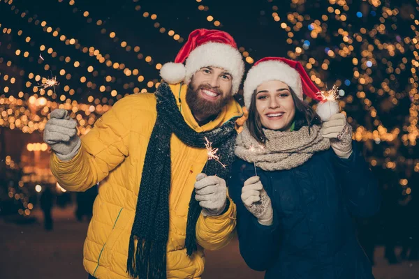Фото пара хлопець леді дві людини на Різдвяному святкуванні в парку, що тримає чарівні блискавки на відкритому повітрі новорічна вечірка в теплих пальто Санта-капелюшках рукавички на вулиці — стокове фото