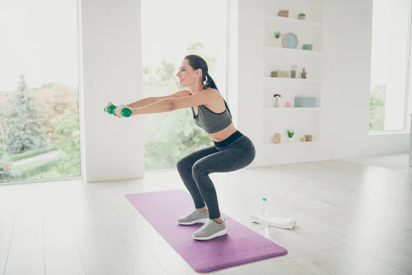 Full-Size-Profil Seite Foto von freudig energische Sportlerin wollen starke Mädchen tun Aerobic sportliche Übung machen Kniebeugen mit ihren Händen halten grüne Hanteln stehen auf lila Matte im Haus wie Turnhalle — Stockfoto