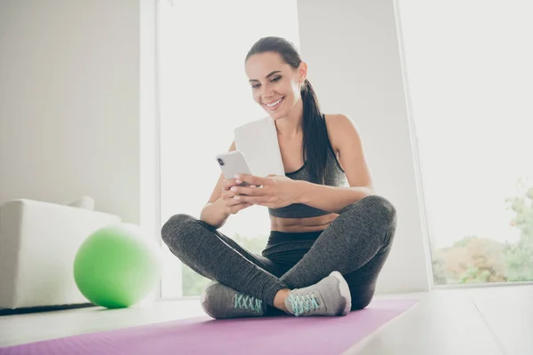 Ganztägiges Foto positiver aktiver, energiegeladener Sportlerin auf lila Matte sitzend Mobiltelefon posten Sportkommentare in gesunden, sportlichen Blogs tragen Turnschuhe graue Hosen im Fitnessstudio wie zu Hause — Stockfoto