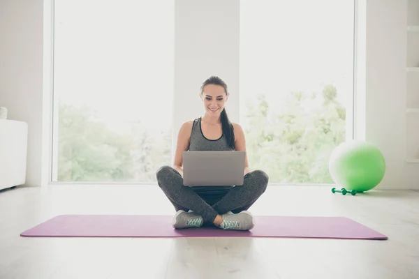 Фотографія повного тіла позитивної спортивної дівчини, яка перетинає ноги, сидить на фіолетовому килимку, використовуючи ноутбук для перегляду фільмів про спорт, що практикують у спортзалі, як будинок у приміщенні — стокове фото