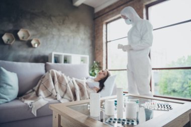 Photo of young patient lady lonely alone lying sofa sick ambulance emergency service visit guy doc virology clinic bring many pills wear uniform hood protection suit visit her flat indoors clipart