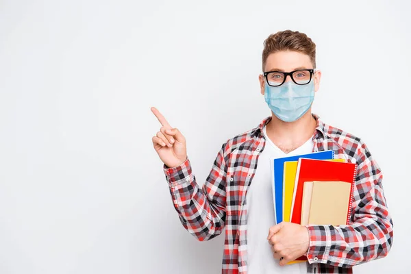 Alegre estudiante nerd celebración de libros y materiales educativos que apuntan a un espacio de copia que muestra la idea de información usar máscara facial de seguridad médica, concepto de educación en línea a distancia remota — Foto de Stock