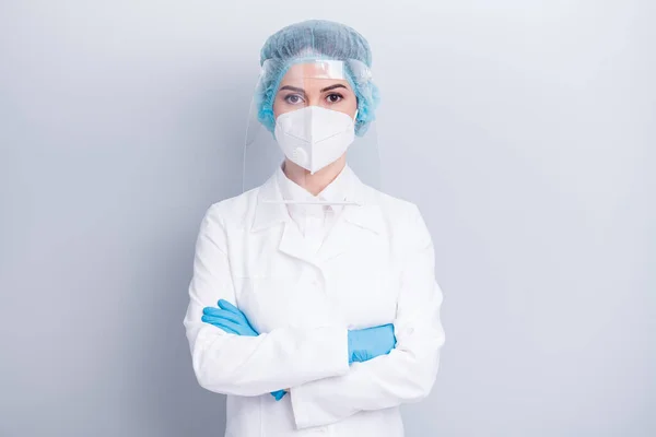 Zdjęcie atrakcyjny pewny wirusolog lekarz ramiona pani skrzyżowane poważne niezawodne zużycie medyczne płaszcz maska twarzy plastik ochrona tarcza chirurgiczne czapka izolowane szare tło — Zdjęcie stockowe
