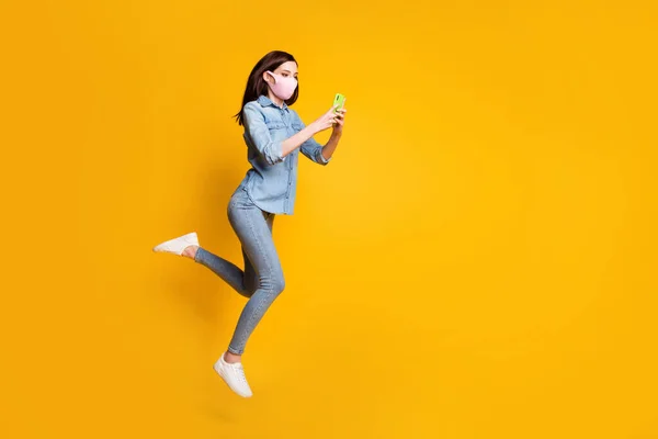 Full storlek foto av vackra flicka hoppa använda smartphone läsa social covid nätverk nyheter bära medicinsk mask denim jeans skjorta isolerad över ljus glans färg bakgrund — Stockfoto