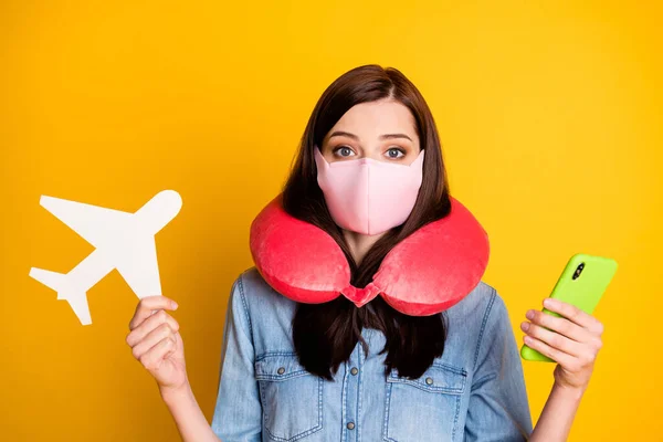 Tıbbi maske takan öğrenci portresi boyun yastığı kullan, akıllı telefon uygulaması kullan, tatil yap, beyaz kağıt uçak giy, kot pantolon giy, sarı arka plan kullan. — Stok fotoğraf