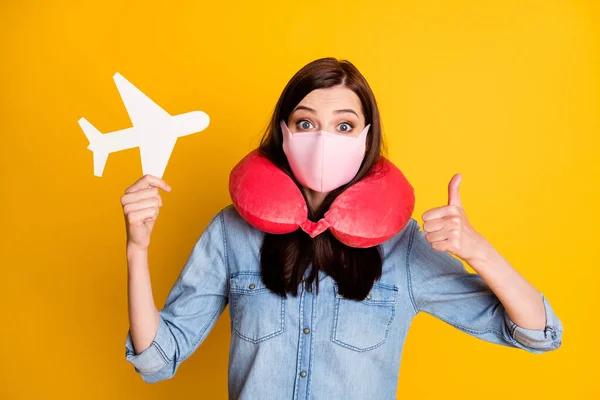 Retrato de bela estudante menina segurar branco papel cartão avião mostrar polegar para cima sinal desgaste viagem macio pescoço almofada médica máscara jeans camisa isolada sobre cor amarela fundo — Fotografia de Stock