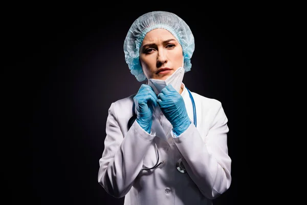 Φωτογραφία της κουρασμένης εξαντλημένης γιατρού κυρία κρατήστε τα χέρια απογειώνεται προστατευτική μάσκα μετά την τελευταία καθυστερημένη λειτουργία αναπνέουν καθαρό αέρα φορούν γάντια εργαστήριο λευκό παλτό χειρουργικό καπάκι απομονωμένο μαύρο χρώμα φόντο — Φωτογραφία Αρχείου