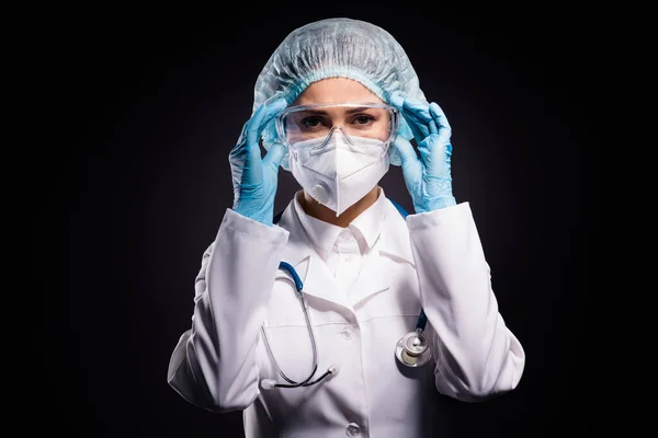Photo of serious lady doc hold arms on protective plastic goggles ready prepare operation wear gloves mask coat facial glasses specs surgical cap isolated black color background — Zdjęcie stockowe