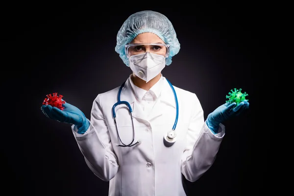 Photo of serious lady doc hold red green covid two bacteria demonstrating difference comparison analysis wear gloves mask coat facial plastic surgical cap isolated black color background
