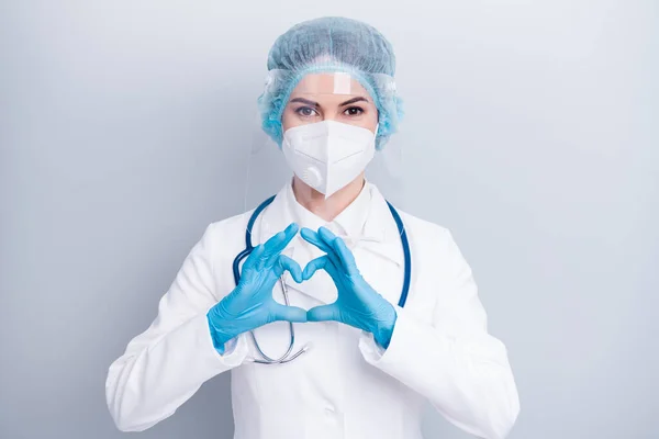 Φωτογραφία της όμορφης κυρίας doc κάνει τα δάχτυλα των χεριών σχήμα καρδιά καλή διάθεση καρδιακή νόσο ευαισθητοποίηση ασφάλεια φθορά γάντια μάσκα παλτό προσώπου πλαστική ασπίδα χειρουργικό καπάκι απομονωμένο γκρι χρώμα φόντο — Φωτογραφία Αρχείου