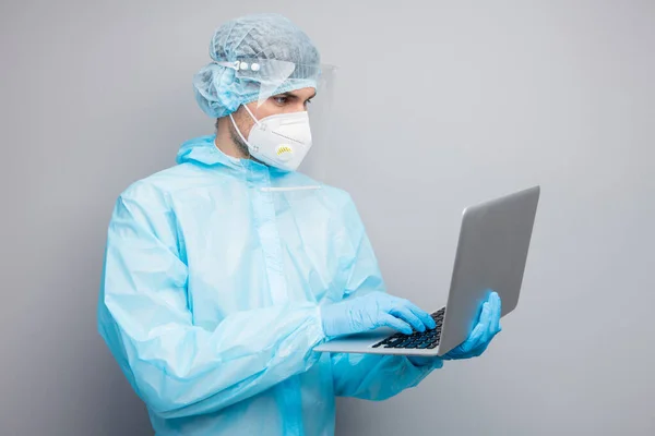 Foto de chico experto doc centro de virología celebrar portátiles hablar pacientes llamada en línea usar máscara respiratoria hazmat azul uniforme traje plástico cara protector escudo aislado gris color fondo — Foto de Stock