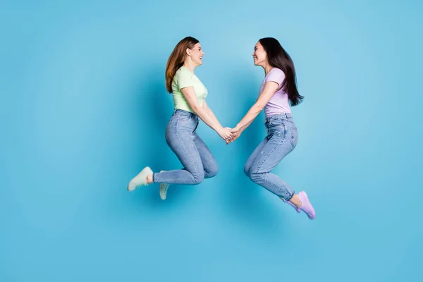 Full body profile photo of two people lesbians couple ladies jump high up hold hands good mood rejoicing happy together wear casual t-shirts jeans footwear isolated blue color background — Stock Photo, Image