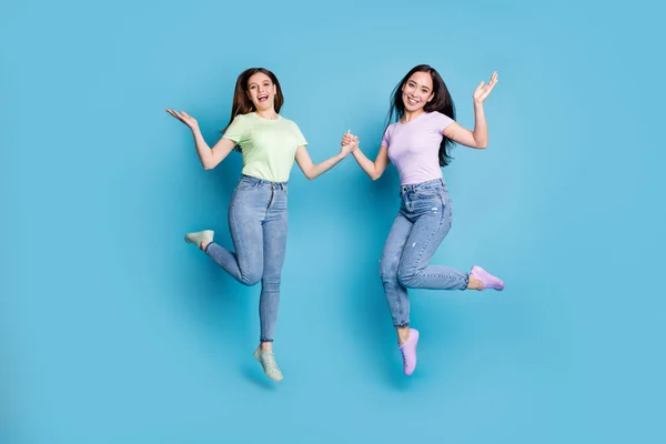 Full length photo of two people lesbians couple ladies jump high up hold hands good mood rejoicing happy together wear casual t-shirts jeans footwear isolated blue color background — Stock Photo, Image