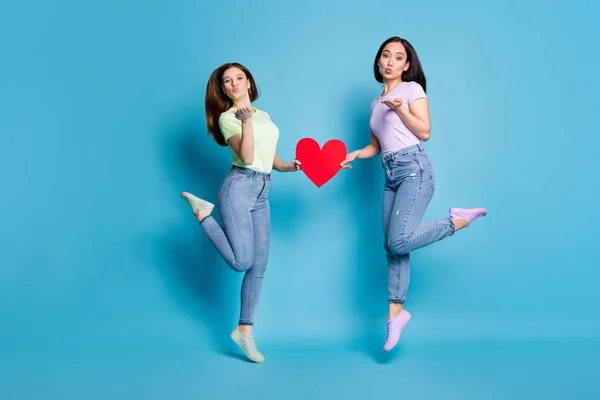 Full size photo of two people lesbians couple ladies jump high up hold big red paper heart valentine day celebration send air kisses wear casual t-shirts jeans isolated blue color background — Stock Photo, Image