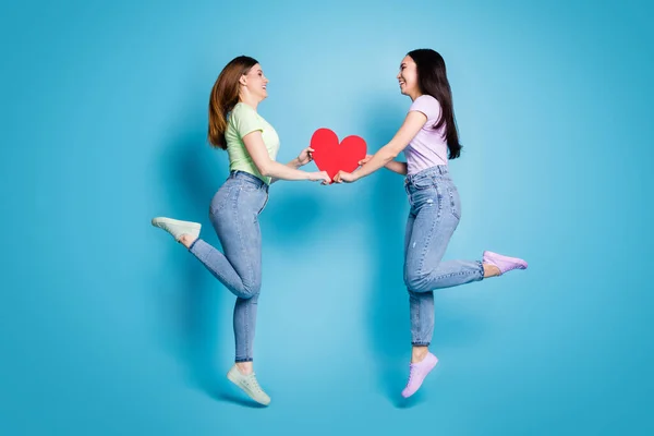 Foto de comprimento total de duas pessoas lésbicas casal senhoras saltar alto segurar grande vermelho papel coração amantes dia dos namorados celebração desgaste casual t-shirts jeans isolado azul cor fundo — Fotografia de Stock