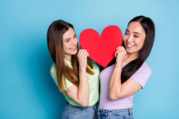 Foto de dos lesbianas divertidas pareja damas romance fecha celebrar papel rojo grande forma del corazón amantes mirar ojos coqueto coqueta astucia desgaste casual camisetas jeans aislado color azul fondo —  Fotos de Stock