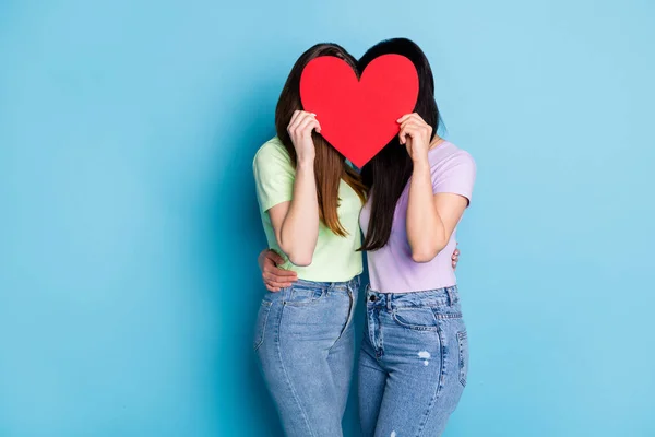 Photo de deux lesbiennes couple jeunes femmes romance date tenant grand papier rouge coeur couverture visages cacher embrasser célébrer anniversaire porter casual t-shirts jeans isolé bleu couleur arrière-plan — Photo