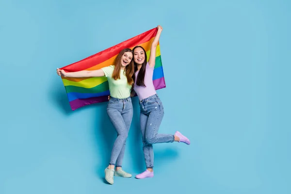 Foto de larga duración de lesbianas encantadoras pareja señoras jóvenes celebran desfile muestran tolerancia todos los matrimonios levantan gay arco iris bandera desgaste casual camisetas jeans aislado color azul fondo — Foto de Stock