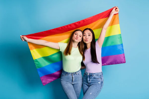 Foto de lesbianas encantadoras pareja jóvenes amantes celebran desfile enviando besos expresan tolerancia todos los matrimonios levantan bandera gay usan camisetas informales jeans aislados color azul fondo — Foto de Stock