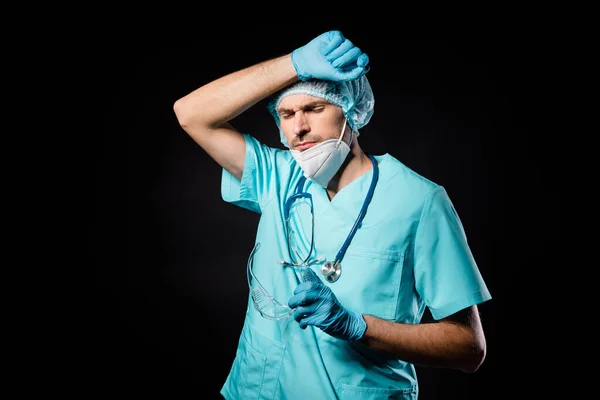 Photo of guy doc professional unhappy hold hand on forehead tiring operation overwhelmed wear mask uniform suit gown plastic facial goggles surgical cap isolated black color background