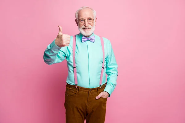 Портрет позитивного веселого старика промоутер шоу большой палец вверх символ рекомендую хороший идеальный рекламный ролик носить бирюзовые брюки фиолетовый галстук бабочка изолированные пастельно-розовый цвет фона — стоковое фото