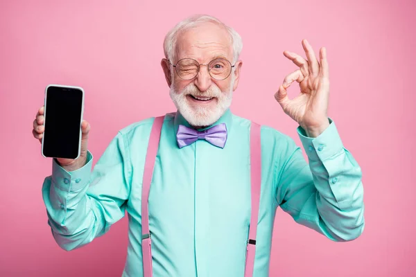 Enerjik, pozitif yaşlı bir adamın portresi yeni bir cep telefonu gösteriyor. Tamam işareti iyi bir modern teknoloji seçmeni tavsiye ederim. Güzel turkuaz gömlek giy, mor papyon, izole edilmiş pastel pembe arka plan. — Stok fotoğraf