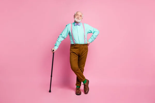 Full length photo of attractive grandpa good mood stand confident lean walking stick wear mint shirt suspenders bow tie brown pants boots green socks isolated pink pastel background — Stock Photo, Image