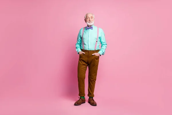 Foto de longitud completa de buen humor atractivo abuelo pie manos confiadas bolsillos llevar camiseta de menta tirantes lazo pantalones marrones botas verdes calcetines aislados rosa pastel fondo —  Fotos de Stock