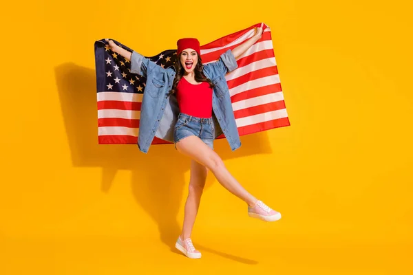 全长合身、精力充沛的街头风格的女孩举着美国国旗，享受着7月4日的庆祝活动，身穿红色头饰牛仔裤背心背心，上衣与亮丽的背景隔离 — 图库照片