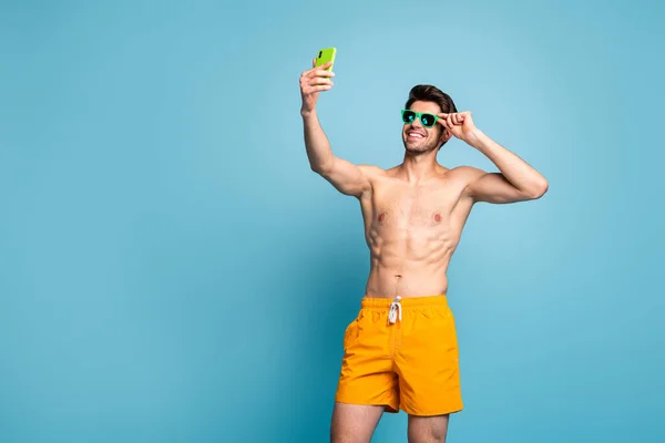 Foto di allegro positivo bell'uomo che regola i suoi occhiali occhio prendendo selfie registrazione video con telefono in pantaloncini gialli isolato colore blu pastello sfondo — Foto Stock