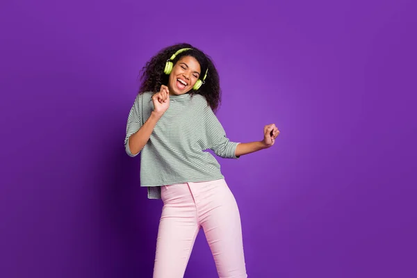 Retrato de menina afro-americana alegre ouvir música alta usar fone de ouvido verde desfrutar cantar música dança sentir regozijar-se usar camisa listrada calças rosa calças isoladas sobre fundo de cor roxa — Fotografia de Stock