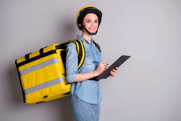 Portret van positieve koerier meisje houden clip board schrijf check lijst genieten leveren pizza thuis dragen thermische rugzak dragen denim outfit helm geïsoleerd over grijze kleur achtergrond — Stockfoto